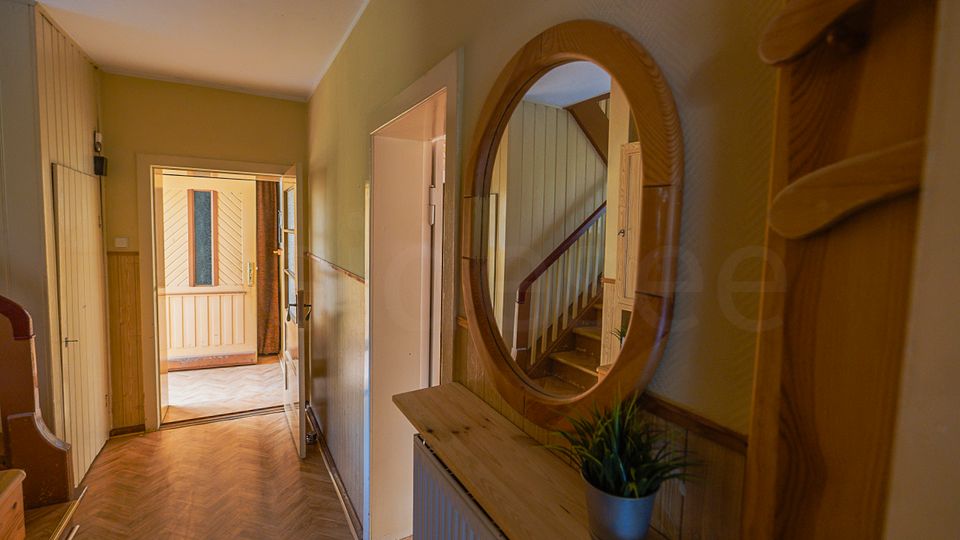 Schmaler Flur in einem Wohnhaus mit Holzpaneelen, Fischgrätparkett und einer Kommode mit rundem Wandspiegel. Am Ende führt eine offene Tür in einen helleren Raum, daneben ist ein Treppenaufgang mit Holzgeländer zu sehen.