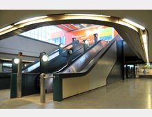 Innenansicht eines U-Bahn- oder Einkaufszentrum-Bereichs mit zwei Rolltreppen und einer Treppe in der Mitte. Im Hintergrund sind große Fenster und farbige Werbeschilder zu sehen, links und rechts schließen sich Geschäfte an.