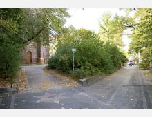 Pflasterweg mit herbstlichem Laub zwischen dichtem Grün, links ein Backsteingebäude mit Holztür, rechts ein Gehweg mit vereinzelten Personen im Hintergrund.
