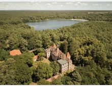 Luftaufnahme eines historischen Gebäudes mit mehreren Türmen und roten Ziegeldächern, umgeben von dichtem Wald. Im Hintergrund befindet sich ein großer See, der von weiteren Waldflächen umgeben ist.