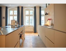 Moderne Küche mit weißen Einbauschränken und Arbeitsflächen aus Edelstahl. Im Hintergrund befinden sich große Fenster mit blauen Vorhängen, die viel Tageslicht hereinlassen.