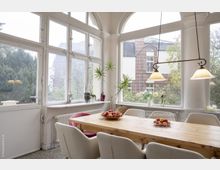 Heller Wintergarten mit großen, weißen Sprossenfenstern und Blick ins Freie, eingerichtet mit einem Holztisch, mehreren Polsterstühlen und einer Hängelampe. Auf dem Tisch stehen Schalen mit Obst, und Pflanzen sowie dekorative Elemente befinden sich auf der Fensterbank.