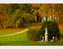 Herbstlicher Park mit einer grünen Wiese, einem geschwungenen Kiesweg und einer steinernen Brunnenfigur auf einem Sockel im Vordergrund. Im Hintergrund stehen dichte Laubbäume mit orangegelbem Herbstlaub, teilweise verdecken sie ein helles Gebäude.