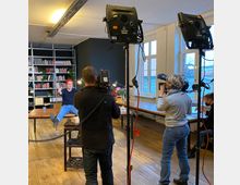 Eine Indoor-Aufnahmeszene in einem Raum mit Holzboden, großen Fenstern und einer Bücherwand im Hintergrund. Eine Person sitzt auf einem Tisch, während zwei Fotografen mit Kamera- und Beleuchtungsausrüstung Aufnahmen machen.