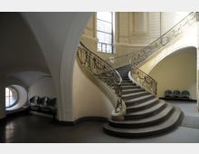 Großzügiges Treppenhaus mit geschwungener Steintreppe und dekorativem Geländer aus Metall. Im Hintergrund befinden sich ein großes Buntglasfenster sowie eine Sitzbank unter einem runden Fensterbogen.