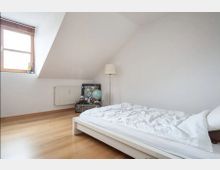 Schlichtes Schlafzimmer mit Dachschräge, einem niedrigen weißen Bett auf Holzboden, einem Fenster mit Holzrahmen und Blick auf eine weiße Wand. Ein Heizkörper, eine Stehlampe und ein niedriger Tisch mit Büchern und einem Globus ergänzen die Einrichtung.