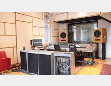 Tonstudio mit einem großen Mischpult, zwei großen Lautsprechern in Holzgehäusen und einem Computermonitor. Die Wände sind mit schallabsorbierenden Paneelen verkleidet, und ein großes Fenster trennt den Regieraum von einem Aufnahmeraum.
