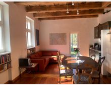 Heller Wohn- und Essbereich mit sichtbaren Holzbalken an der Decke, Holzboden und großen Fenstern entlang der linken Wand. Im Raum stehen ein ovaler Esstisch mit Stühlen, ein rotes Ecksofa und rechts eine kompakte Küchenzeile; hinten führt eine Glastür nach draußen.