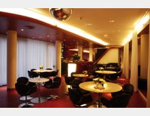 Moderner Innenraum einer Lounge mit roten Böden und einer abgerundeten Decke in warmem Licht. Mehrere weiße Tische mit schwarzen Stühlen, dazu Dekorationen wie weiße Blumen auf den Tischen; im Hintergrund eine Bar mit beleuchtetem Tresen und Sitzbereichen.