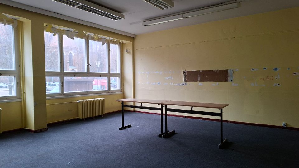 Leerer, älterer Büroraum mit gelb gestrichenen Wänden, Teppichboden und einer langen Tischreihe in der Mitte. Große Fensterfront mit Heizkörper darunter und Deckenleuchten; an der rechten Wand sind entfernte Verkleidungen bzw. abgeblätterte Stellen sichtbar.