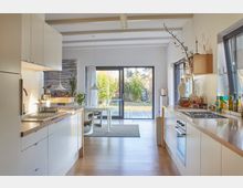 Moderne Küche mit weißen Schränken, Holzarbeitsflächen und einem großen Fenster im Hintergrund, das den Blick auf einen Garten mit Rasen und Bäumen freigibt. Zentral steht ein Esstisch unter zwei hängenden Lampen, und durch eine große Glasschiebetür fällt viel Tageslicht ein.