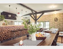 Gemütliches Café mit rustikaler Einrichtung, inklusive Holztischen und -bänken mit blau-weiß karierten Tischdecken. Eine Theke mit Holzscheiben-Verkleidung, hängenden Lampen und dekorativen Pflanzen prägen den Raum; im Hintergrund eine grüne Tapete mit pinkfarbenem Muster.