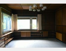 Ein schlichtes Interieur mit hölzernen Wänden und Einbaumöbeln, großen Fenstern mit beigen Vorhängen, und einer modernen Deckenleuchte. Der Raum hat einen hellen Teppichboden, und durch die Fenster fällt Tageslicht mit Blick auf eine Baumlandschaft.