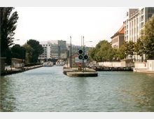 Stadtkanal mit klarem Wasser, flankiert von modernen Gebäuden und Bäumen; im Vordergrund eine kleine Insel mit Schiffssignallichtern, im Hintergrund eine Brücke und ein Schiff.