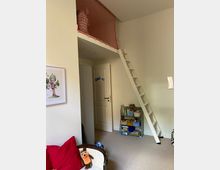 Zimmer in einem Wohnhaus mit Hochbett-Mezzanine, weißer Leiter und rotem Sicherheitsnetz. Im Vordergrund ein Sessel mit rotem Kissen und Stofftier, daneben ein offenes Regal mit Büchern und Spielzeug, sowie eine weiße Tür im Hintergrund.