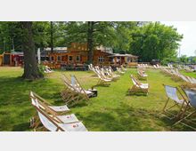 Grüne Außenanlage mit Holzgebäuden im Hintergrund, umgeben von Bäumen. Auf der Rasenfläche stehen zahlreiche Liegestühle und Holztische, die auf einen gastronomischen Bereich hinweisen.