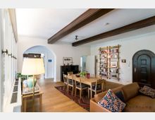 Wohnzimmer mit Holzboden, einem Holztisch und mehreren Stühlen. Links ein großes Fenster, rechts ein Bücherregal an der Wand und eine dunkle, rundbogige Tür. Im Hintergrund ein Klavier und ein Durchgang mit gewölbtem Türrahmen.