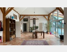 Ein heller Wohnraum mit sichtbaren Holzbalken, einem Esstisch aus Holz und großen Glasfenstern auf der rechten Seite. Der Raum verfügt über Terrakottafliesen, eine Bücherwand links sowie Zimmerpflanzen und Türen im Hintergrund.