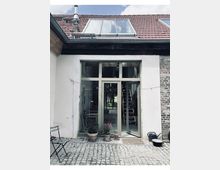 Innenhofansicht eines Hauses mit rotem Ziegeldach und Dachfenster sowie großer, bodentiefer Glasfront mit Rahmen und Oberlicht. Vor dem Eingang liegen Kopfsteinpflaster, mehrere Blumentöpfe und eine Gießkanne; seitlich sind ein kleiner Tisch und eine Ziegelwand zu sehen.