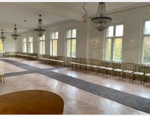 Ein heller, großer Saal mit hohen Fenstern, poliertem Holzboden und stilvollen Kronleuchtern an der Decke. Entlang der Wände stehen goldene Stühle mit hellen Polstern in einer Reihe, während ein grauer Läufer diagonal über den Boden verläuft.