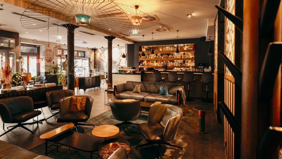 Offener, modern gestalteter Bar- und Loungebereich mit Ledersesseln, Sofas und niedrigen Couchtischen auf Fliesenboden. Im Hintergrund eine lange Theke mit beleuchtetem Flaschenregal, schwarze Säulen und auffällige, fächerförmige Deckenleuchten in warmem Licht.