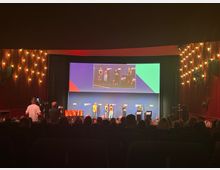 Ein Innenraum eines Theaters oder Kinos mit einem beleuchteten Bühnenbereich, auf dem mehrere Personen stehen. Im Hintergrund ist eine große Leinwand mit einer Präsentation zu sehen, und das Publikum sitzt im abgedunkelten Saal.
