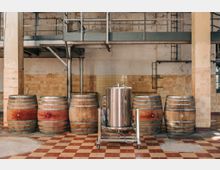 Innenraum einer Weinkellerei mit mehreren Holzfässern entlang einer Wand sowie einem metallischen Gärtank auf Rollen in der Mitte. Der Raum hat eine helle, geflieste Wand, eine industrielle Metallkonstruktion im oberen Bereich und einen Boden mit rot-gelben Kacheln.