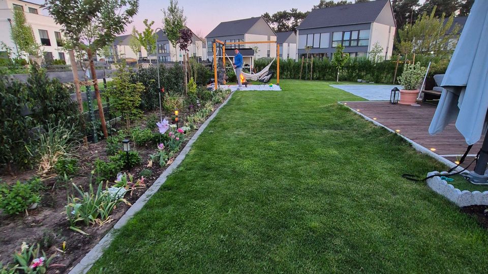 Großer, gepflegter Hausgarten mit breitem Rasenstreifen, links mit Beeten und jungen Bäumen bepflanzt und rechts mit Holzterrasse sowie Sonnenschirm. Im Hintergrund stehen moderne Wohnhäuser, und in der Mitte des Gartens befindet sich ein Holzgestell mit Hängematte.