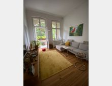 Wohnzimmer mit Holzboden, einer grauen Ecksofa mit gelbem Kissen, einem kleinen Glastisch und einem gelben Teppich. Im Hintergrund befinden sich große Fenster und eine Balkontür, die zu einem begrünten Außenbereich führen. Ein farbenfrohes Bild hängt an der Wand.