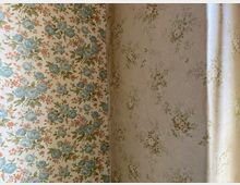 Nahaufnahme einer Raumecke mit zwei unterschiedlich gemusterten Blumentapeten, links mit blau-rosa Blumen und rechts mit dezenteren, kleinen Blumenmotiven. Rechts fällt weiches Tageslicht entlang der Wand in den Raum.