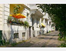 Straßenansicht eines Altbauwohnhauses mit weißen Fassaden, kleinen Balkonen und gelben Sonnenschirmen. Vor dem Gebäude stehen Fahrräder, Pflanzen zieren die Balkone, und ein gepflasterter Gehweg verläuft entlang der Straße, die von Bäumen gesäumt ist.