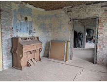 Ein Innenraum mit rohen, teils unverputzten Backsteinwänden und abblätternder gelb-blauer Wandfarbe. Im Vordergrund steht eine antike Holzorgel; durch eine offene Tür sind weitere Räume mit unvollendeter Einrichtung, darunter ein Schaufenstermannequin, sichtbar.