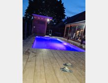 Ein beleuchteter Außenpool mit lila Licht, umgeben von einer Holzterrasse, mit einem kleinen Holzhäuschen links und einer verglasten Hausfassade rechts. Im Hintergrund stehen Bäume, und der Himmel zeigt eine abendliche Dämmerung.