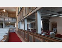 Innenansicht einer historischen Kirche mit hölzernen Emporen und verzierten Paneelen. Im Vordergrund sind Holzbänke mit einem roten Teppich in der Mitte, im Hintergrund eine Orgel und Wandtafeln mit Text und Dekorationen sichtbar.