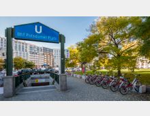 Eingang zur U-Bahn-Station „Bhf Potsdamer Platz“ in Berlin mit blauem Schild, umgeben von modernen Gebäuden und herbstlichen Bäumen. Rechts stehen mehrere Parkräder, links führt eine Treppe zur U-Bahn hinunter.