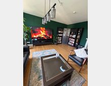 Wohnzimmer mit dunkelgrünen Wänden und Holzdielenboden, mit großem Fernseher auf niedrigem Sideboard, der ein Kaminfeuer zeigt. In der Ecke stehen schwarze Bücherregale mit Schrankelement, davor ein Teppich und ein Glastisch; darüber hängt eine Deckenleuchte mit mehreren Glaszylindern.
