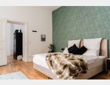 Schlafzimmer mit einem großen Bett, weißen Kissen, einer braunen Felldecke und einer grünen Tapete mit Pflanzenmuster. Im Hintergrund führt eine offene Tür in einen angrenzenden Raum mit hölzernem Bodenbelag und einem schwarzen Schrank mit grüner Zimmerpflanze oben drauf.