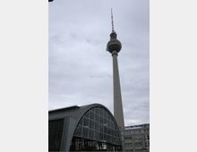 Fernsehturm in Berlin vor bewölktem Himmel, im Vordergrund ein halbkreisförmiges Glasdach eines Bahnhofsgebäudes. Rechts daneben sind Teile eines rechteckigen Bürogebäudes erkennbar.