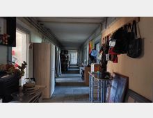 Ein langer, schmaler Flur mit einfacher Beleuchtung und Betonboden. Links stehen Schränke und kleinere Möbelstücke, rechts hängen Taschen an einer Wand, und mehrere Regale und Gegenstände sind entlang der Länge des Gangs verteilt.