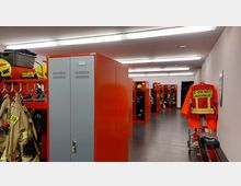 Umkleideraum einer Feuerwehrstation mit roten Spinden und offenen Fächern, die Schutzkleidung und Helme enthalten. An der Wand hängt eine leuchtend orangefarbene Dienstjacke mit der Aufschrift „C-Dienst Luckenwalde“, der Boden besteht aus dunklen Fliesen.