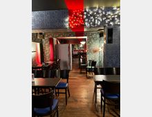 Ein gemütliches, dimm beleuchtetes Restaurant oder Café mit Holzboden, dunkelblauen gepolsterten Stühlen und floral gemusterten Wänden in Blau und Rot. Lichterketten schmücken die Decke, während eine Bar im Hintergrund zu sehen ist.