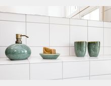 Weiße, geflieste Badezimmerablage unter einem Fenster mit grüner Keramik-Seifenspenderflasche, Seifenschale mit Stückseife und zwei passenden Bechern. Die rechteckigen Wand- und Ablagefliesen prägen das helle, minimalistische Ambiente.