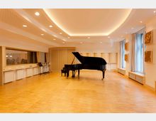 Ein heller Aufnahmeraum mit Holzboden und schalldämpfenden Elementen an den Wänden. In der Mitte steht ein schwarzer Flügel, daneben befinden sich Mikrofonständer; große Fenster lassen Tageslicht hinein.