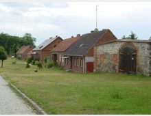 Reihe von Back- und Natursteinhäusern mit Satteldächern in einem ländlichen Dorf; im Vordergrund eine gepflasterte Straße und eine grüne Wiese mit einzelnen Bäumen und Sträuchern.