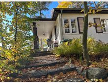Einfamilienhaus mit weißer Fassade und Fenstern mit Holzläden, umgeben von Herbstlaub und Farnen. Ein gepflasterter und mit Blättern bedeckter Weg führt durch einen Garten zu einer überdachten Terrasse.