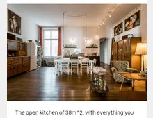 Offene Wohnküche mit Holzboden und einer Kombination aus modernen und Vintage-Möbeln. Die Küche im Hintergrund verfügt über eine Arbeitsfläche und Regale, während im Vordergrund ein Esstisch mit Stühlen sowie ein kleiner Wohnbereich mit Sessel, Couchtisch und einer Stehlampe zu sehen sind.