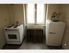 Kleine Küche mit einem alten weißen Elektroherd, einem weißen Kühlschrank der Marke Bosch und einem Fenster mit Heizkörper. Vor dem Fenster stehen zwei alte Holzkisten, auf dem Kühlschrank liegen Geschirr und Küchenutensilien.