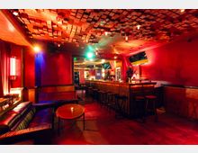 Innenraum einer Bar mit roter Wandgestaltung, langer hölzerner Theke mit Barhockern und gedämpfter, farbiger Beleuchtung. Markant sind die Decke mit akustischen Holzpaneelen, eine Sitzecke mit Sofa und runden Tischen sowie Wandmonitore und Spiegel hinter der Bar.