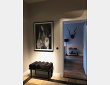 Ein Flur mit cremefarbenen Wänden, einem gerahmten Schwarz-Weiß-Fotoporträt an der Wand und einer schwarzen gepolsterten Sitzbank darunter. Im Hintergrund ist ein angrenzender Raum mit Parkettboden, einem Geweih an der Wand und einem kleinen Servierwagen zu sehen.