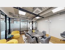 Besprechungsraum mit hellen Wänden, einem runden Tisch mit grauen Stühlen und zwei gelben Sesseln. Der Raum verfügt über eine Glaswand mit satiniertem Muster, eine sichtbare Deckeninstallation und Holzboden.